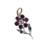 An antique-style floral pendant charm featuring a silver-toned metal setting with a central clear round stone surrounded by six deep pinkish-red round stones, with two green accent stones on leaf-shaped settings below, all on a plain white background.